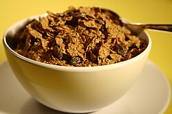 A white ceramic bowl of flake cereal with added raisins that is placed on top of a matching saucer.