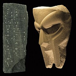 A stone or concrete rendition of the modern MF Doom mask, facing a stone with abstract symbols etched into it.