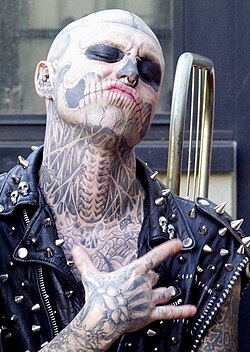 A man covered in skeleton-like tattoos on his face, neck, and hands, wearing a spiky leather vest.