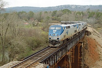 Two monocoque locomotives leading eight single-level cars across a small bridge