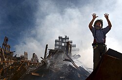 A firefighter stands on rubble, holding up 10 fingers to request assistance