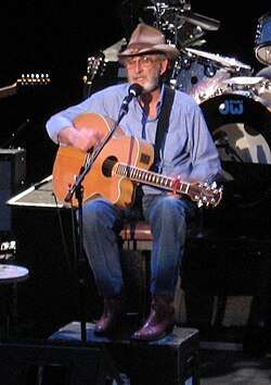 Don Williams, seated and playing an acoustic guitar while singing into a microphone