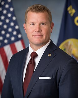 Official Senate portrait of Sheehy smiling in front of the U.S. and Montana flags, wearing a black suit, white shirt, and floral red tie.