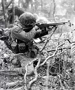 U.S. Marine Corps infantryman kneeling in underbrush and firing a Browning Automatic Rifle while aiming into area out of frame.