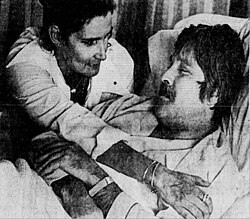 A man with a mustache lying in a hospital bed. A woman comforts him.