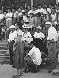 A crowd of people gathered at a film location shoot: in the background, slightly out of focus are many adults and children, some standing, some sitting on stone steps; in the left foreground is a young Japanese woman, Hara, in a white blouse and dark dress, with camera crew behind her; a middle-aged Japanese man, Ozu, in dark pants, white shirt and floppy hat stands at far right foreground.