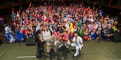 Group shot taken from the stage, facing the audience. The celebrities are crouched down with their backs to the audience while the audience is mostly standing to pose for the photo. Many people in the audience are wearing cosplay.