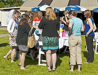 A group of women gathered around a table in an outdoor setting, dressed in a mix of casual and dressy styles. At the left of the image is a woman wearing a veil on the back of her head.