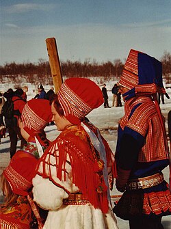 People wearing red and blue winter garments with patterned trim, tall fabric hats, wide belts, and shawls stand outdoors in snow during a spring gathering.