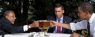 three men in neckties and suit jackets sit around a white table outdoors while clinking frothy mugs of pale-amber beer together in a toast over two silver bowls on a silver tray, one containing salted pretzels.
