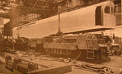 A picture of a locomotive under construction. The cuboid box containing cabs and boiler is being lowered onto the two bogies by a crane inside a locomotive works. Various components are scattered in the foreground.