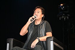 Low angle photograph of a mustached Japanese man in a black outfit sitting in a chair speaking into a microphone.