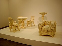 Four cafe chairs and a table, a high table, and an upholstered chair are wrapped in fabric and rope.