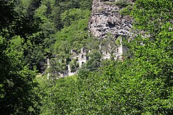 An overgrown, multi-story stone building in ruins. It lies in the mountains, surrounded by forest.
