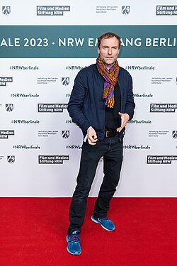 Mark Waschke wearing light blue running shoes, dark jeans, a dark shirt under a dark blue open jacket, and a colourful scarf, standing in front of a 2023 Berlinale festival stage backdrop, looking toward camera but not directly at it, with slight grin on his face