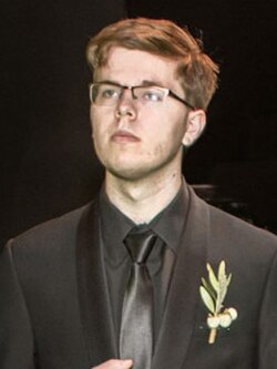 Formally dressed, 20-year-old, White man wearing a side-part in black tuxedo, black button down shirt, and black tie with glasses standing still