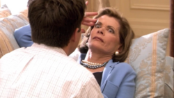 An elderly women with her hand on her forehead stares with her eyes wide open at a man sitting across from her