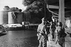 men n quayside looking across water at burning tanks