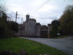 Stone building with slit windows and battlements. Foreground is road with grass verges.