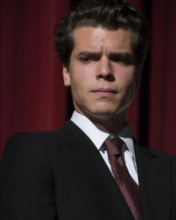 Photograph of Gus Halper as Erik Menendez in a suit, staring pensively at what can be presumed to be the ground.