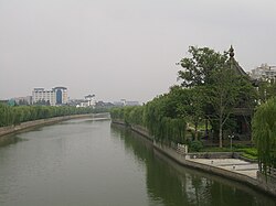 A canal scene: on the left bank stands a small pavilion surrounded by willows and other trees; along the right bank runs a row of willows, with high-rise buildings in the background