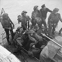 A black and white photograph showing soldiers wearing combat equipment sitting in a small watercraft beside a pier