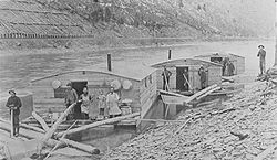 Three Arks for a log drive on Pine Creek, in Lycoming or Tioga County, Pennsylvania. The left ark was for cooking and dining, the middle ark was the sleeping quarters and the right ark was for the horses. The arks were built for just one log drive and then sold for their lumber. The line of the Jersey Shore, Pine Creek and Buffalo Railway can be seen on the eastern shore: the mountainside behind it is nearly bare of trees from clearcutting.