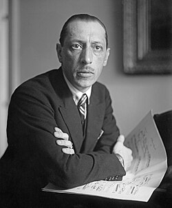 Black and white photo of Stravinsky resting his arms atop a piano, a score under his hands