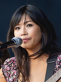 Beabadoobee behind the mic, playing guitar, made-up and wearing a floral blouse.