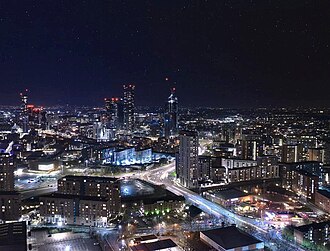 A skyline of a city against a blue sky and tall hills in the background. There are around nine skyscrapers distributed fairly evenly across the skyline with many cranes also visible. In the foreground there is a river with trees on both banks.