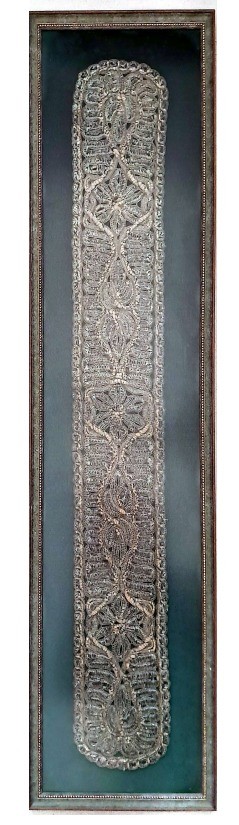 A close-up photograph of a framed, long rectangular silver lace neckband for a prayer shawl, featuring intricate symmetrical patterns of interlaced metallic threads on a dark background.