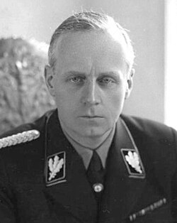 Portrait of a middle-aged man with short grey hair and a stern expression. He wears a dark military uniform, with a swastika on one arm. He is seated with his hands on a table with several papers on it, holding a pen.