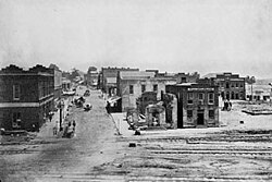 About a dozen one- and two-story buildings, several of which are damaged, line a dirt road that intersects with three railroad tracks in the foreground