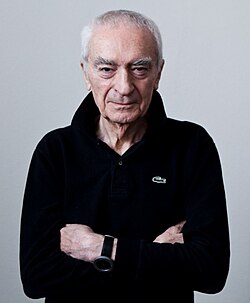 Elderly Vignelli pictured facing the camera, arms crossed, wearing a black, long-sleeved Izod polo shirt with an upturned collar.