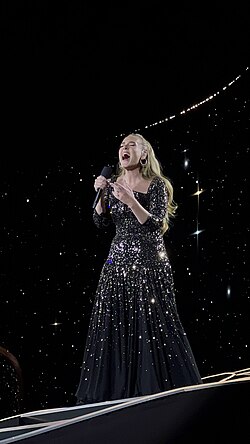 Adele sings into her microphone, dressed in a black gown ornamented with mirror shards