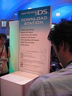 A kiosk with a large sign that fades vertically downward from light blue to white. The words "Nintendo DS Download Station" are above a list of demo names, which includes "Electroplankton (E3 demo)".