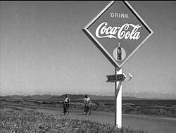 A young Japanese man and woman, both in casual clothes, are riding bicycles over a paved road in the near background of the image; mountains are visible in the far distance. In the foreground of the image, at the edge of the road, is a diamond-shaped Coca-Cola sign, below which is an arrow upon which is written the name (in English) of a beach.