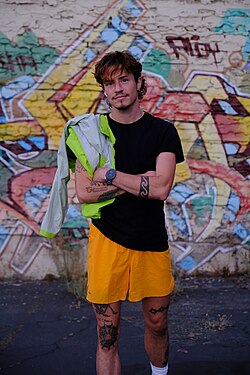 Calamia stands before a graffiti-covered wall wearing a black t-shirt, orange shorts and a black wristwatch. A lime green jacket is slung over his shoulder and his arms are crossed over his chest. Black tattoos are visible on his forearms and legs.
