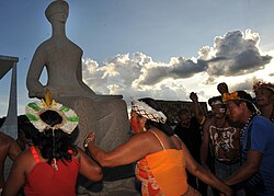 Indigenous people holding hands, two women in the foreground, surround a statue that represents Justice