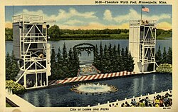 Color postcard depicting a swimming pool surrounded by a seating area, and a stage with diving platforms on both ends