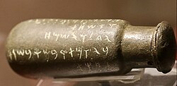 A bronze bottle on its side with text in the Phoenician alphabet "𐤅𐤀𐤔𐤇𐤕 / 𐤉𐤂𐤋 / 𐤅𐤉𐤔𐤌𐤇 /𐤁𐤉𐤅𐤌𐤕 𐤓𐤁𐤌 𐤅𐤁𐤔𐤍𐤕"