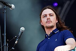 Against a black concert background punctuated by a hazy white light, a man in a blue shirt stands next to a pair of microphones.