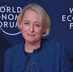A woman with short blonde hair wearing a blue jacket speaks at a podium in front of a World Economic Forum backdrop