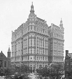 A black-and-white photograph shows a grand, ornate multi-story Beaux-Arts style building with elaborate facades, towers, and decorative details.
