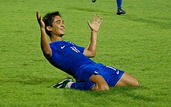 Chhetri on his knees in a blue football shirt and blue football shorts. His arms are outstretched and he is smiling after just scoring a goal.