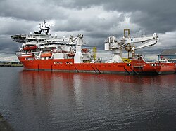 A red hulled ship seen from the port quarter at a low angle. It has heavy lifting gear on the quarterdeck and a helipad over the forecastle.