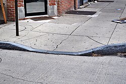 A granite curb cut without tactile paving. A signpost and the foot of a brick building are visible in the background, along with detritus on the sidewalk.
