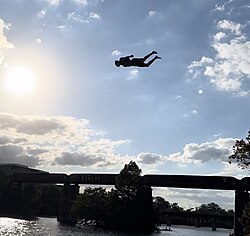 Jackwagonboy mid-flight on a classic death dive off a bridge, body locked in the signature face-down, flat position as the water rushes up to meet him.