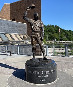 Statue of Roberto Clemente wearing a baseball uniform and holding up his cap. Circular base says his name, 1934-1972, and Goya.