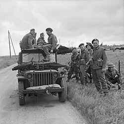 A black-and-white photograph of a jeep with soldiers sitting on top and standing beside it. The soldiers sitting on the jeep are three German soldiers and one British soldier who is interrogating the Germans. On the bonnet of the jeep is small motorcycle, while in the background is a Horsa glider